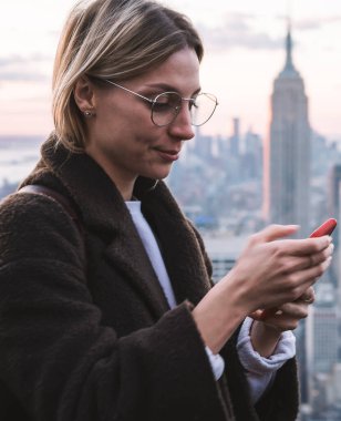 Sarışın bayan turist, tatillerde New York 'u ziyaret ederken Empire State binasını manzaralı açık bir güvertede beklerken cep telefonu kullanıyor. Hippi kız seyahat blogu yapıyor