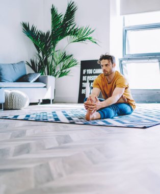 Genç adam yoga eğitimine başlamadan önce modern apartman dairesinde şık bir iç mekanla halıda oturuyordu. Spor amacı olan ve evinde antrenman yaparken bacağını esneten motivasyonlu bir hippi.