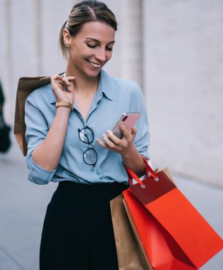 Sosyal ağda akıllı telefondan mesajlar okuyan olumlu hipster kızı şehir ortamında ayakta duruyor, satış ve indirimli mağazaları ziyaret ettikten sonra cep telefonu kullanarak gülümsüyor.