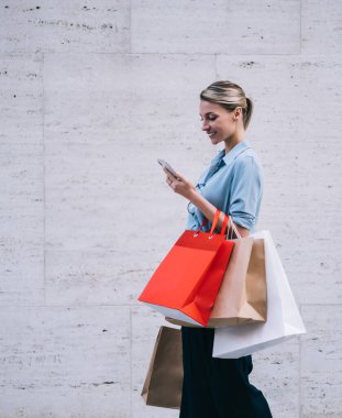 Elinde kağıt paketler olan mutlu bir kadın alışverişçi, marka adı için reklam metni için fotokopi alanının yakınında yürüyor, cep telefonunda GPS uygulaması kullanan pozitif bir kadın.