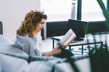 Modern dizüstü bilgisayarın üstünde oturmuş, şık iç dekorasyonlu bir şekilde kitap okuyan gülümseyen genç bir kadın. Evde boş zamanlarında edebiyat entrikalarından hoşlanan pozitif hipster kız.
