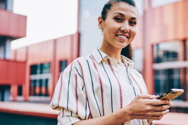 Modern akıllı telefondan sosyal ağlara medya indirirken kameraya gülümseyen pozitif Japon genç kadın portresi açık havada umumi 4G internet kullanarak.