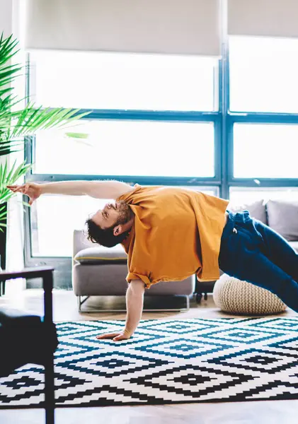 Experienced male lover of yoga in sportive wear stretching hand standing in yoga pose during morning training at home interior.Professional sportive man practising meditation in modern apartment