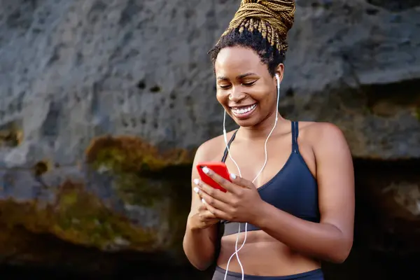 Happy dark skinned female jogger happy from cool music on radio for morning workout outdoors, cheerful excited woman listening audio from player in headphones connected to cellular before training