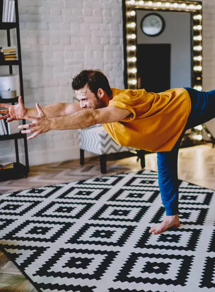 Concentrated young man raising hands hands forward standing on one leg on cozy carpet during morning workout in modern apartment with stylish interior.Motivated hipster guy doing yoga exercises