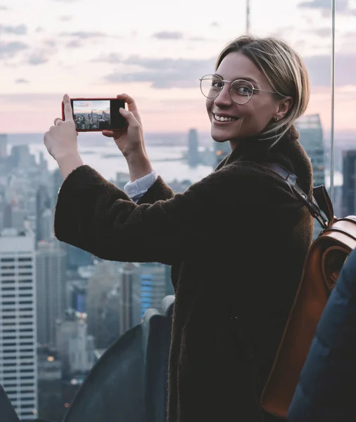 Happy carefree female tourist taking photo on smartphone camera while visiting famous Observation deck with scenery view of New York landmarks. Hipster girl enjoying her ravel to USA during vacations
