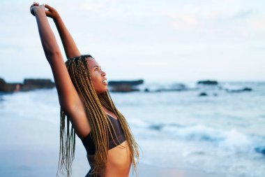 Koyu tenli sporcu germe ve okyanus yakınlarındaki kıyı şeridi nde sabah egzersiz sırasında enerji eğitimi yapıyor, çekici genç kadın koşucu açık havada koşu başlamadan önce esneklik egzersizleri yapıyor