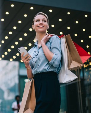 İyi görünümlü beyaz bir müşteri cep telefonuyla alışveriş merkezinin yanında duruyor. Kara Cuma 'da alışveriş yapıyor. Kafkasyalı, kağıt torbalı neşeli bir müşteri akıllı telefon cihazında internet bağlantısı kullanıyor.