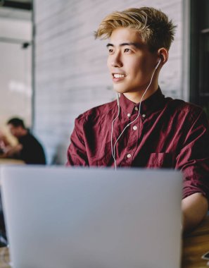 Başarılı Asyalı erkek serbest çalışan, modern dizüstü bilgisayar, eğlence ve keyif kavramı ile uzaktan çalışırken elektronik ekipmanlarla favori müzik listesini dinlerken başka yöne bakıyor.