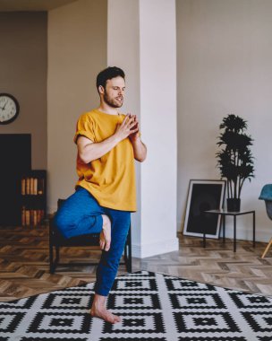 Düşünceli genç adam namaste 'de el ele tutuşurken rahat bir halının üzerinde duruyor ve modern apartman dairesinde sabah meditasyonu sırasında şık iç mimarisi olan ahengi düşünüyor.