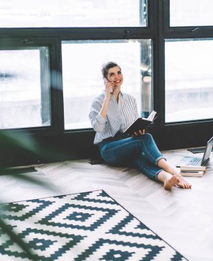 Dijital akıllı telefon cihazından genç bir kadın arıyor ve arkadaşıyla buluşmayı planlamak için konuşuyor modern apartman dairesinde not defteriyle otururken şık iç mimarisi ve büyük pencereleri var.