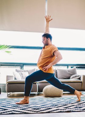 Spor yapan genç bir adamın sabah evde çalışırken el ele tutuşması. Hippi adam yoga eğitiminden önce ısınıyor. Havalı halı ve ahenk ve sağlıklı zindeliğin keyfini çıkarıyor.