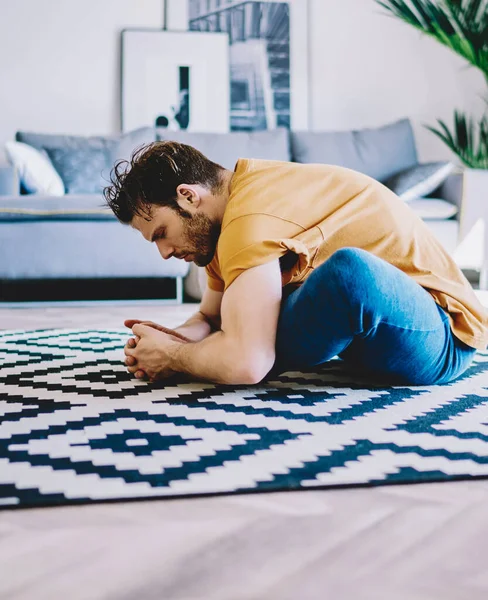 Side view of young man sitting in lotus pose on carpet and doing stretching exercises in modern apartment.Motivated hipster girl practicing yoga and healthy lifestyle at home interior