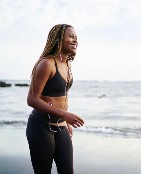 Cheerful african female runner dressed in sport top listening favourite music songs in modern earphones during sport workout walking on sea coastline of tropical island, concept of healthy lifestyle
