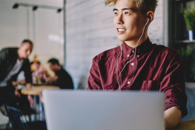 Başarılı Asyalı erkek serbest çalışan, modern dizüstü bilgisayar, eğlence ve keyif kavramı ile uzaktan çalışırken elektronik ekipmanlarla favori müzik listesini dinlerken başka yöne bakıyor.