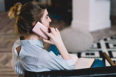 Modern bir dairede oturmuş akıllı telefondan gülümseyen ve konuşan neşeli genç bir kadın. Daire içinde cep telefonuyla konuşan neşeli bir hipster blogcu.