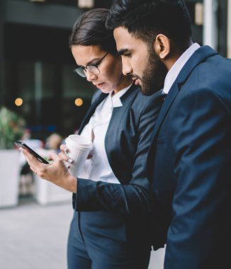 Konsantre olmuş yetişkin erkek ve kadın iş ortakları, finans sitesinde, kahve molası sırasında internette gezinmek için 4G kablosuz internet bağlantısı olan dünya kurumsal anlaşmasıyla ilgili haberleri okuyorlar.