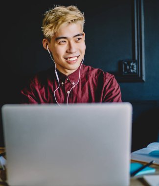 Olumlu gülümseyen Asyalı erkek öğrenci modern dizüstü bilgisayarla sohbet etmekten ve elektronik kulaklıkla güzel sesleri dinlemek için yeni müzik uygulamaları kullanmaktan keyif alıyor.