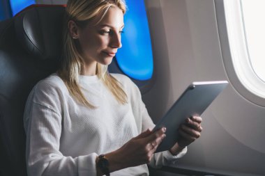 Genç beyaz kadın yolcu modern elektrokromik teknolojiyle geniş kontrollü pencerenin yanında geniş koltukta rahat bir uçuş yapıyor. Turist kadın gemide kablosuz bağlantı kullanıyor