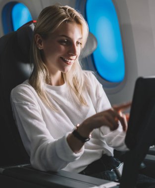 Gülümseyen çekici bayan yolcu rahat uçuş sırasında TV cihazından izlemek için film arıyor. Beyaz tenli genç kadın gezgin uçakta dokunmatik ekran eğlence izleme sisteminin keyfini çıkarıyor.