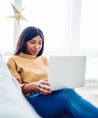 Pozitif Afro-Amerikalı kadın web sitesi sayfasına göz atıp rahat koltukta internet üzerinden alışveriş yapmak için malzeme seçiyor. Koyu tenli genç hippi kız ev içi bloglarda dizüstü bilgisayar kullanıyor.