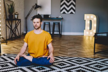 Yoğunlaşmış, gözleri kapalı, uyumun ve vücut şeklinin tadını çıkaran genç bir adam. Sabah meditasyonu sırasında Lotus pozisyonunda otururken modern apartmanın iç kısımlarında dinleniyor.