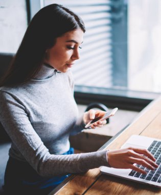 Düşünceli iş kadını 20s kahve dükkanında oturan web mağazasında alışveriş için dizüstü bilgisayar kullanırken modern akıllı telefon uygulaması üzerinden ödeme yaparken, teknoloji ve iletişim kavramı