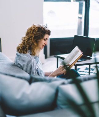 Modern dizüstü bilgisayarın üstünde oturmuş, şık iç dekorasyonlu bir şekilde kitap okuyan gülümseyen genç bir kadın. Evde boş zamanlarında edebiyat entrikalarından hoşlanan pozitif hipster kız.