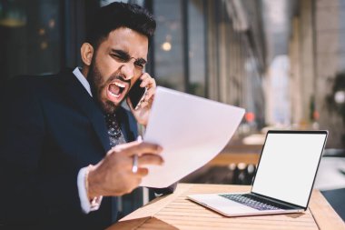 Öfkeli İspanyol erkek şirket müdürü akıllı telefon görüşmeleri sırasında internet reklamınız için netbook 'la masada oturuyor ve bağırıyor.