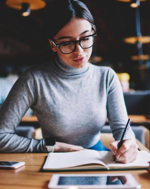 Düşünceli hipster kız 20s kafeteryada oturan, gözlük kadın pensive öğrenci üniversite kampüsü içinde ders kitabı ile eğitim notu okuma ve planları hakkında düşünme, iş kadını notlar yapma