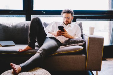 Gözlüklü, ciddi bir beyaz adam cep telefonu numarasıyla ev içi koltukta yatıyor, bin yıllık hipster adam akıllı telefondan posta ve mesajları kontrol ediyor ve online dağıtım numaralarını arıyor.