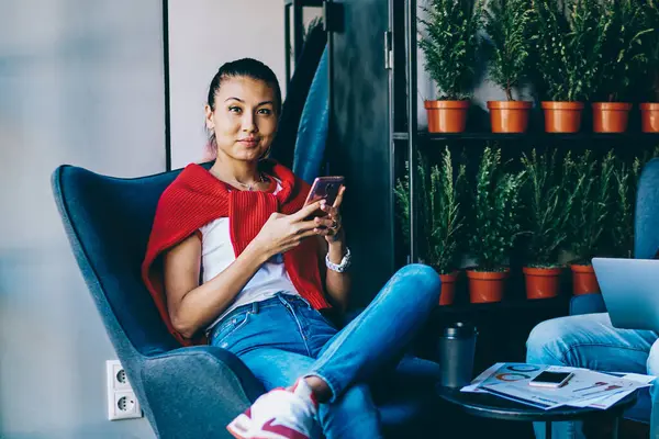 Portrait of positive female blogger sitting in comfortable chair in cafeteria with cozy interior and holding smartphone gadget while blonde chinese male friend near ignoring live communication