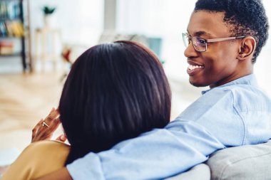 Gülümseyen Afro-Amerikan erkeği kız arkadaşına sevgiyle bakıyor oturma odasında dinlenirken konuşuyor, hafta sonları evdeki kanepede oturup dinlenen koyu tenli çiftin dikiz aynasından bakıyor.