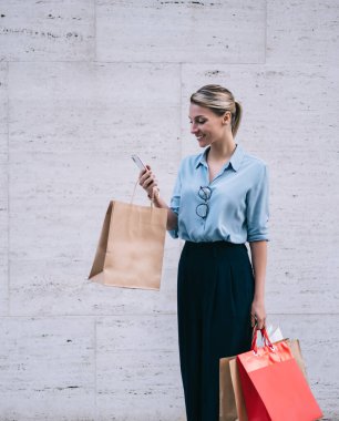 Kara Cuma 'da alışveriş gününden sonra online sohbet için akıllı telefon cihazını kullanan mutlu hippi kız, reklam için fotokopi alanının yanında duran kağıt torbalı beyaz müşteri.
