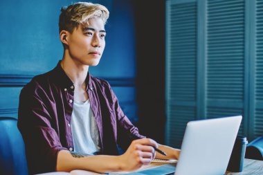 Düşünceli Asyalı hippi öğrenci geliştirme için yaratıcı fikirler üretiyor. Kendi başlangıç projesini düşünüyor. Masa başında dizüstü bilgisayarla oturuyor.