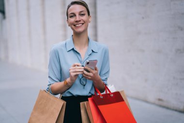 Heyecanlı kadın alıcının yarım boy portresi kağıt paketlerle tanıtım alanında duruyor ve kameraya gülümsüyor modaya uygun giyinmiş mutlu hippi kız elinde modern cep telefonu cihazıyla dışarıda bekliyor.