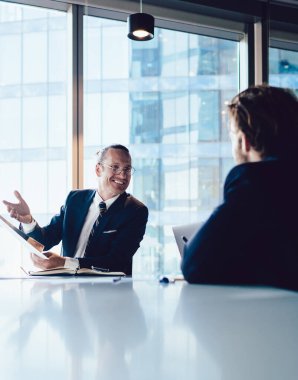 Şirket meslektaşları ofis içinde toplantı masasında oturup gülümseyen erkek yöneticilerle fikir alışverişinde bulunuyorlar. Yaratıcı stratejileri ve beyin fırtınalarını paylaşan bir grup işçi