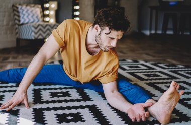 Evdeki yoga egzersizleri sırasında halının üzerinde oturup esneklik kazanması için motive olmuş genç bir adam. Deneyimli hippi adam apartmanda bacak esnetiyor ve sağlıklı yaşam tarzını geliştiriyor.