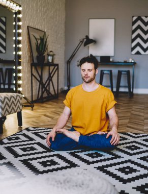 Yoğunlaşmış, gözleri kapalı, uyumun ve vücut şeklinin tadını çıkaran genç bir adam. Sabah meditasyonu sırasında Lotus pozisyonunda otururken modern apartmanın iç kısımlarında dinleniyor.