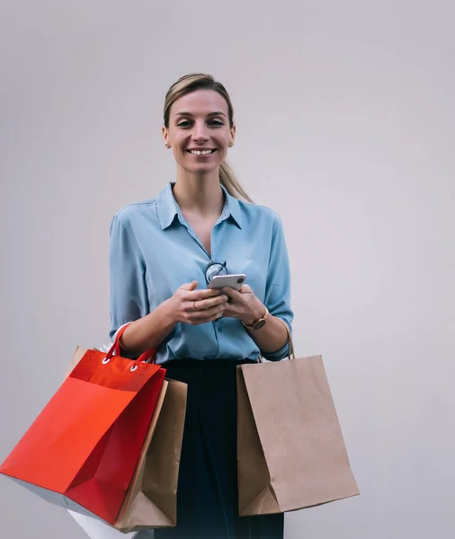 Half length portrait of positive woman in stylish outfit holding cellular phone and paper bags in hands and smiling at camera, cheerful millennial shopaholic standing on promotional background