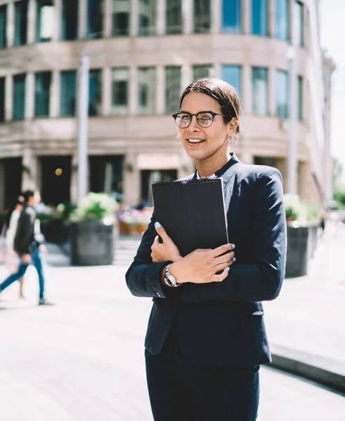 Half length portrait of confident positive office worker holding folder and smiling at camera during work break in business district, happy caucasian employee dressed in elegant suit feeling good