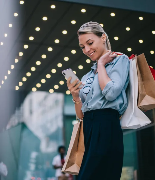 Stylish woman with paper bags in hands spending time near shopping center and reading incoming message on smartphone device, happy hipster girl customer with packages dialing number on mobile phone