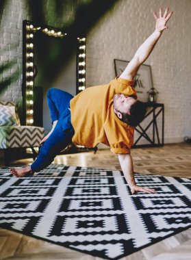 Yoganın profesyonel erkek aşığı, sabah antrenmanı sırasında modern dairede şık bir halı üzerinde poz veriyor. Sağlıklı yaşam tarzını desteklemek için spor egzersizleri yapan deneyimli genç adam.