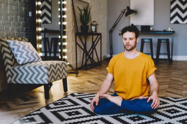 Yoğunlaşmış, gözleri kapalı, uyumun ve vücut şeklinin tadını çıkaran genç bir adam. Sabah meditasyonu sırasında Lotus pozisyonunda otururken modern apartmanın iç kısımlarında dinleniyor.