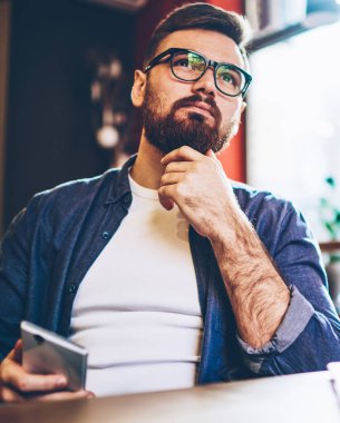 Düşünceli erkek blogcu gündelik kıyafetlerle kendi web sayfası için kafa yoruyor. Kafenin içinde boş boş boş oturuyor, tefekkür eden beyaz bir adamla buluşmayı düşünüyor. Arkadaşını içeride beklerken.