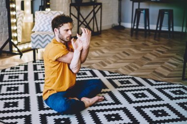 Asana duruşunda elini uzatan genç adam modern apartmandaki rahat halının üzerinde Lotus 'ta oturuyor. 20' li yaşlardaki hippi çocuk evde iç mekan eğitimi sırasında meditasyon yapıyordu.