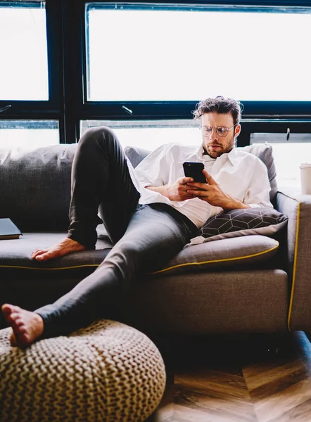 Serious caucasian man in spectacles dialing number on mobile phone lying on sofa in home interior, millennial hipster guy checking mail and messages on smartphone searching number of online delivery