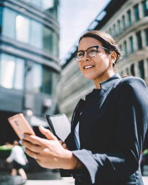 Görme düzeltmesi için optik gözlüklü beyaz kadın. Başka tarafa bakıyor ve gülümsüyor. Resmi takım elbise giymiş, elinde akıllı telefon cihazıyla aramasını bekliyor.