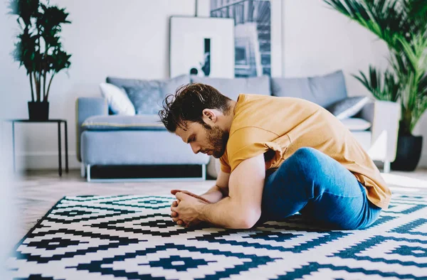 Side view of young man sitting in lotus pose on carpet and doing stretching exercises in modern apartment.Motivated hipster girl practicing yoga and healthy lifestyle at home interior