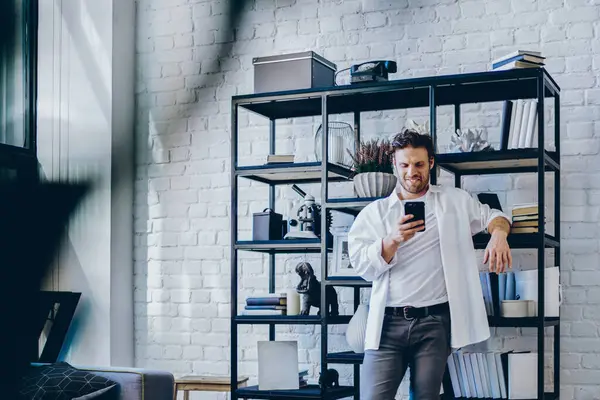 Positive smiling man in casual wear standing in modern designed living room in apartment with mobile phone in hand, hipster guy sending text messages on smartphone using app and 4G connection